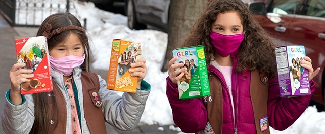 Two masked Girl Scouts outdoors holding up Girl Scout Cookie boxes